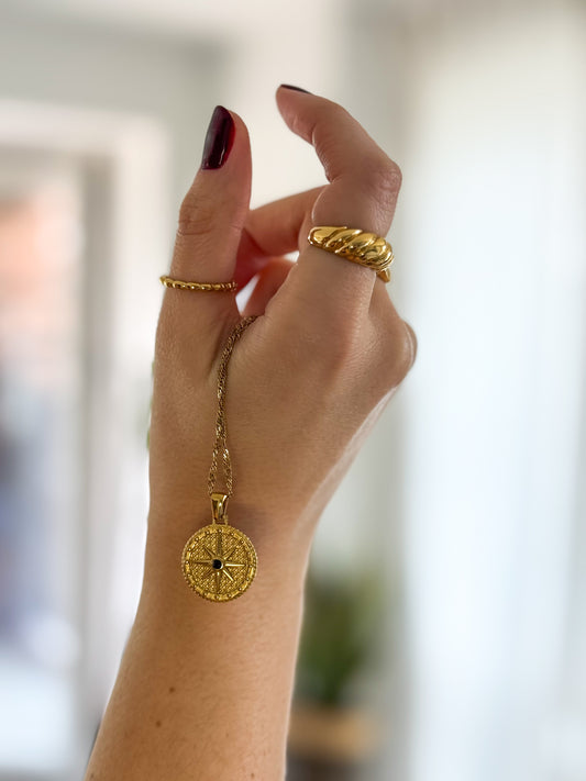 Hand wearing a gold ring and gold necklace with a circular pendant against a blurred background