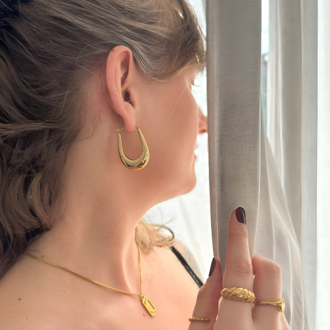 Person wearing gold hoop earrings and rings, looking out a window.